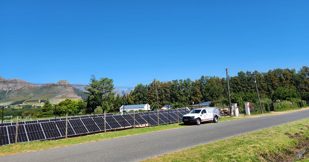 Long row of solar panels installed along rural roadside with white service truck, mountains and vineyards visible